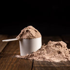 Skopa med chokladfärgat proteinpulver och vit burk, med pulver på träyta